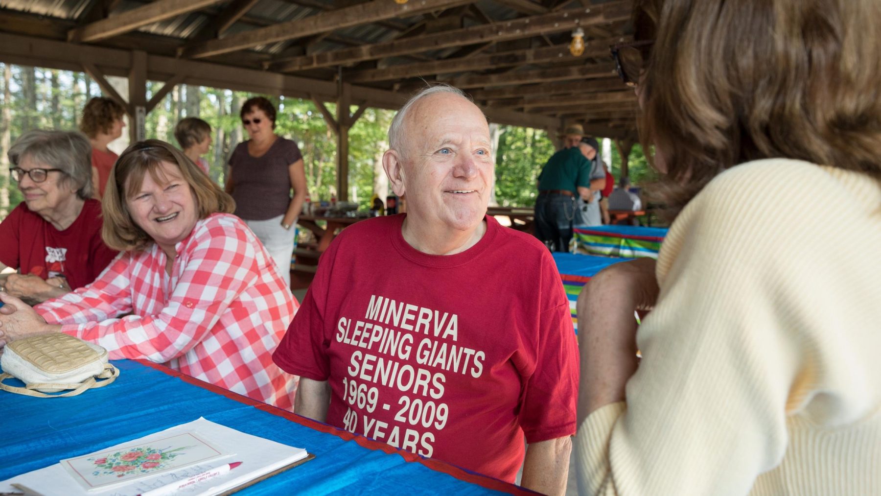 a gray-haired man wearing a shirt that says, "Minerva Sleeping Giants" talking with two women